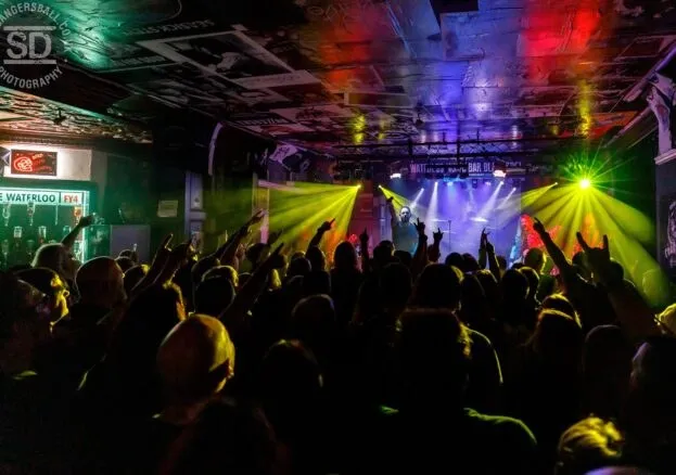 Packed gig crowd inside Waterloo Music Bar with stage lighting and a lit "Waterloo FY4" bar sign in the background.