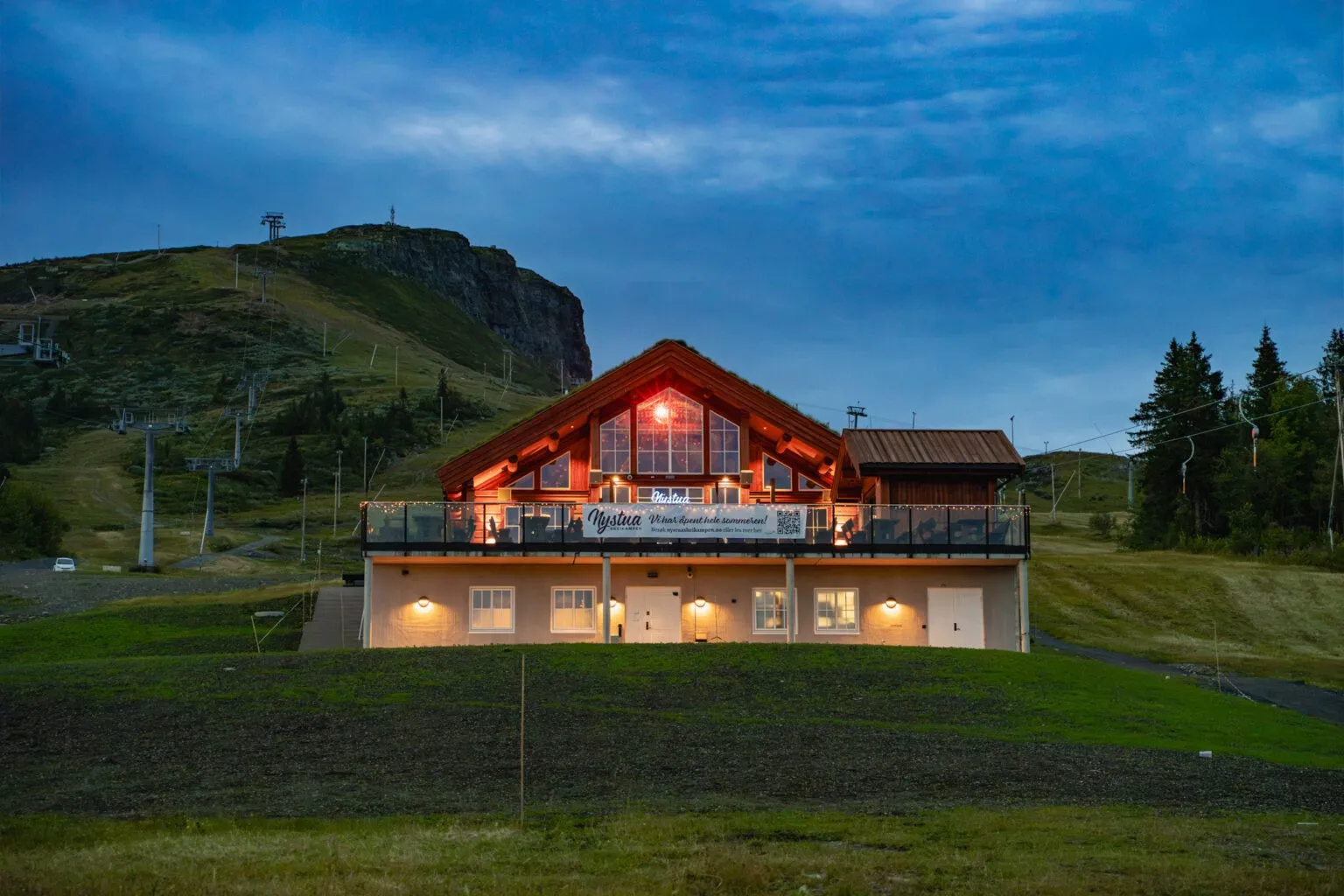 Nystua's sod-roofed timber lodge at dusk on the Skeikampen ski slope, with a lit "Nystua" sign in the window and a banner reading "Nystua — vi har åpent hele sommeren!" on the terrace rail