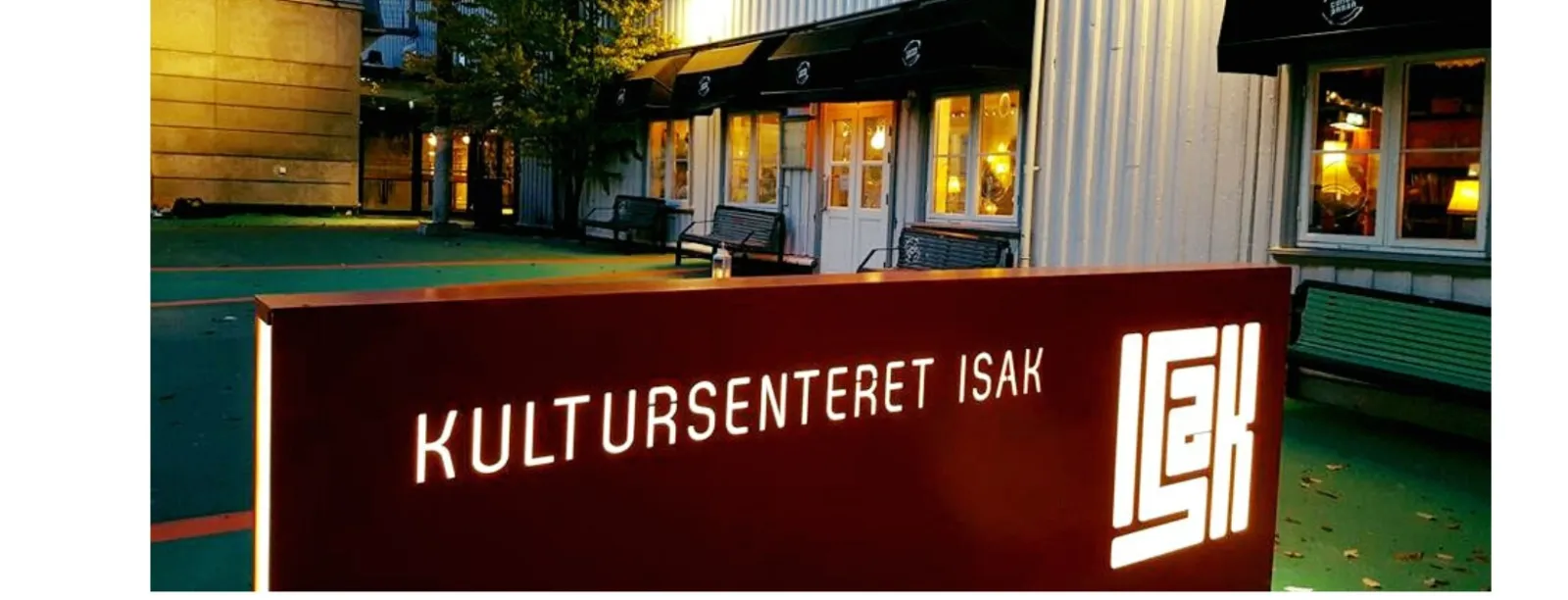 Kultursenteret ISAK at dusk: a white weatherboarded building with black window awnings and warmly lit windows, seen across its green courtyard, with a large illuminated dark red sign in the foreground reading "KULTURSENTERET ISAK" beside the ISAK monogram.