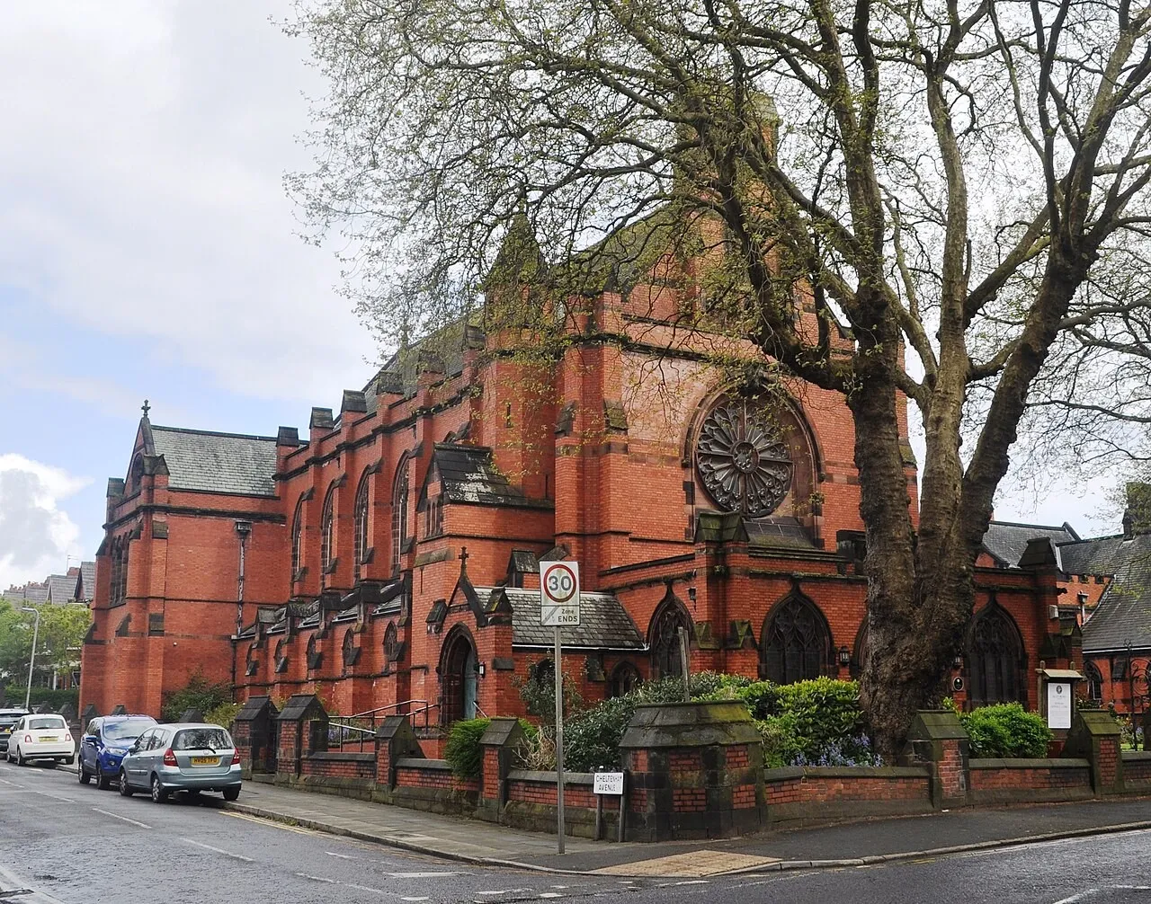 The church from the pavement opposite, the rose window gable and the porch framed between bare trees, with parked cars along the kerb and a 20 mph sign in the foreground.