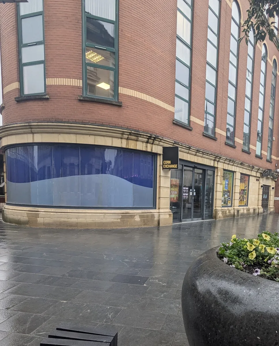 Entrance of the Corn Exchange on Newport High Street, glass doors under a black Corn Exchange sign in a stone-faced ground floor with gig posters in the windows, red-brick storeys above